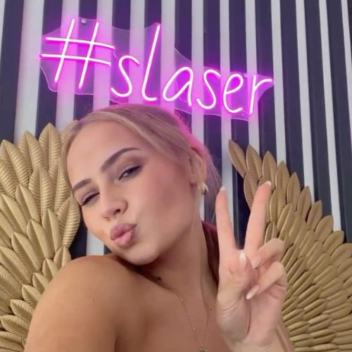 Frau steht vor einem pinken Neonschild mit der Aufschrift #slaser und zeigt mit der Hand das Peace-Zeichen